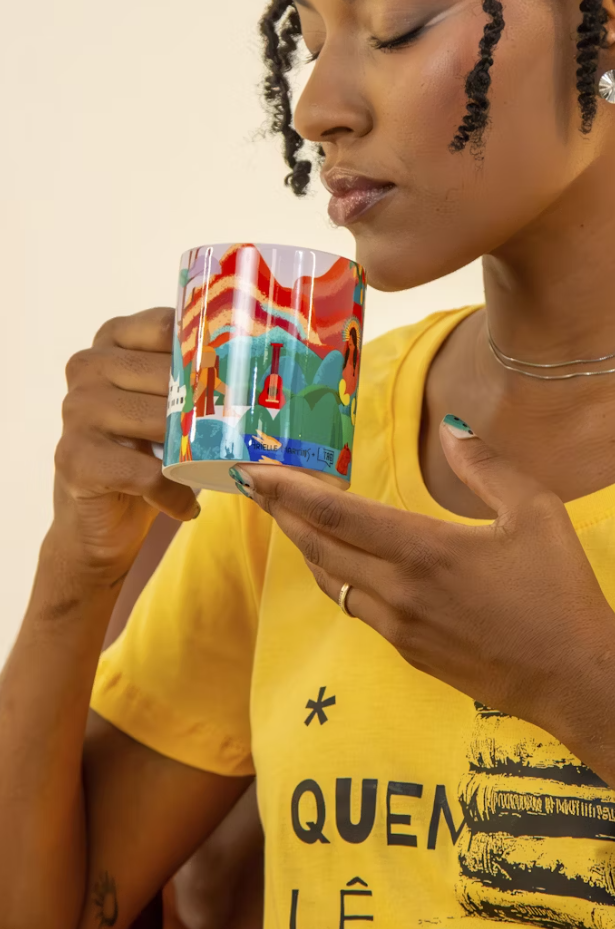 Modelo segura uma caneca chico rei próximo da boca, é uma caneca com bastante cores