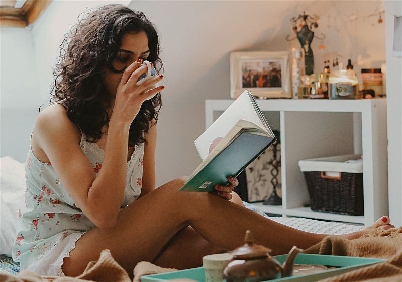 Mulher sentada em cama tomando algo em xícara e lendo livro